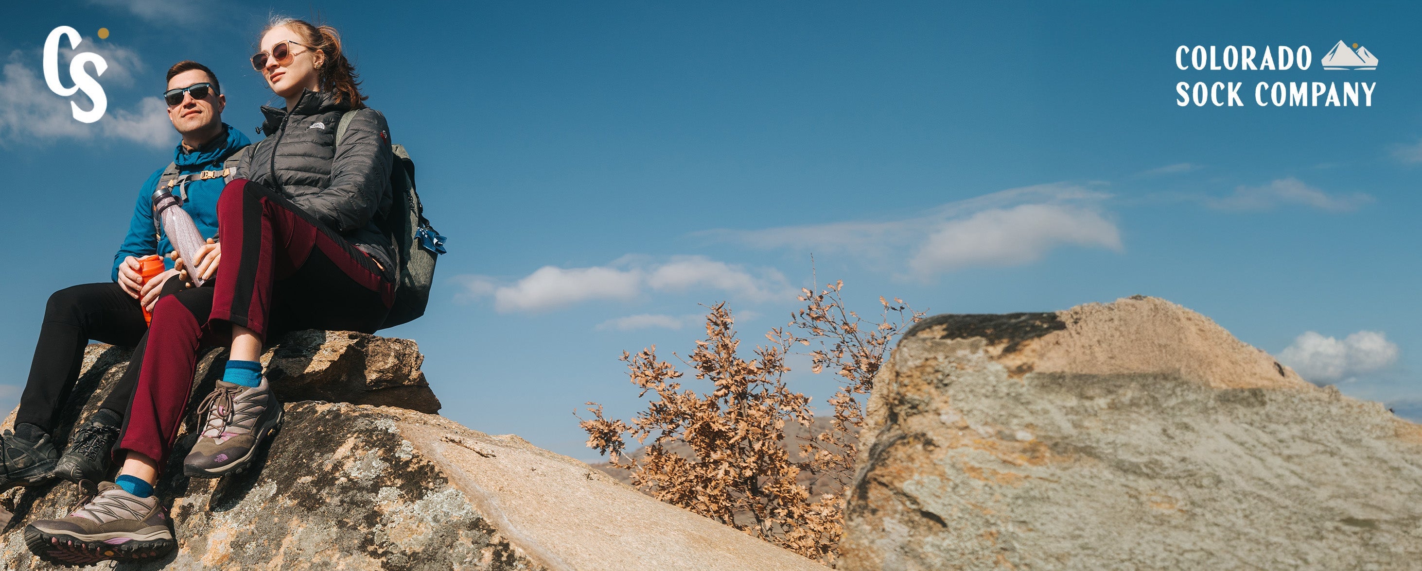 Two people sitting on a rocky outcrop with a clear blue sky and 'Colorado Sock Company' branding.