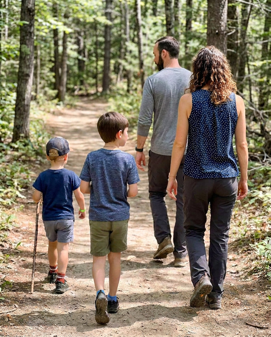 A family of four hiking on a dirt trail through a dense forest, photographed from behind. The mother wears a sleeveless navy blue top with small white dots and dark gray pants, the father wears a gray long-sleeve shirt and dark pants, and the two children wear blue and green outfits.