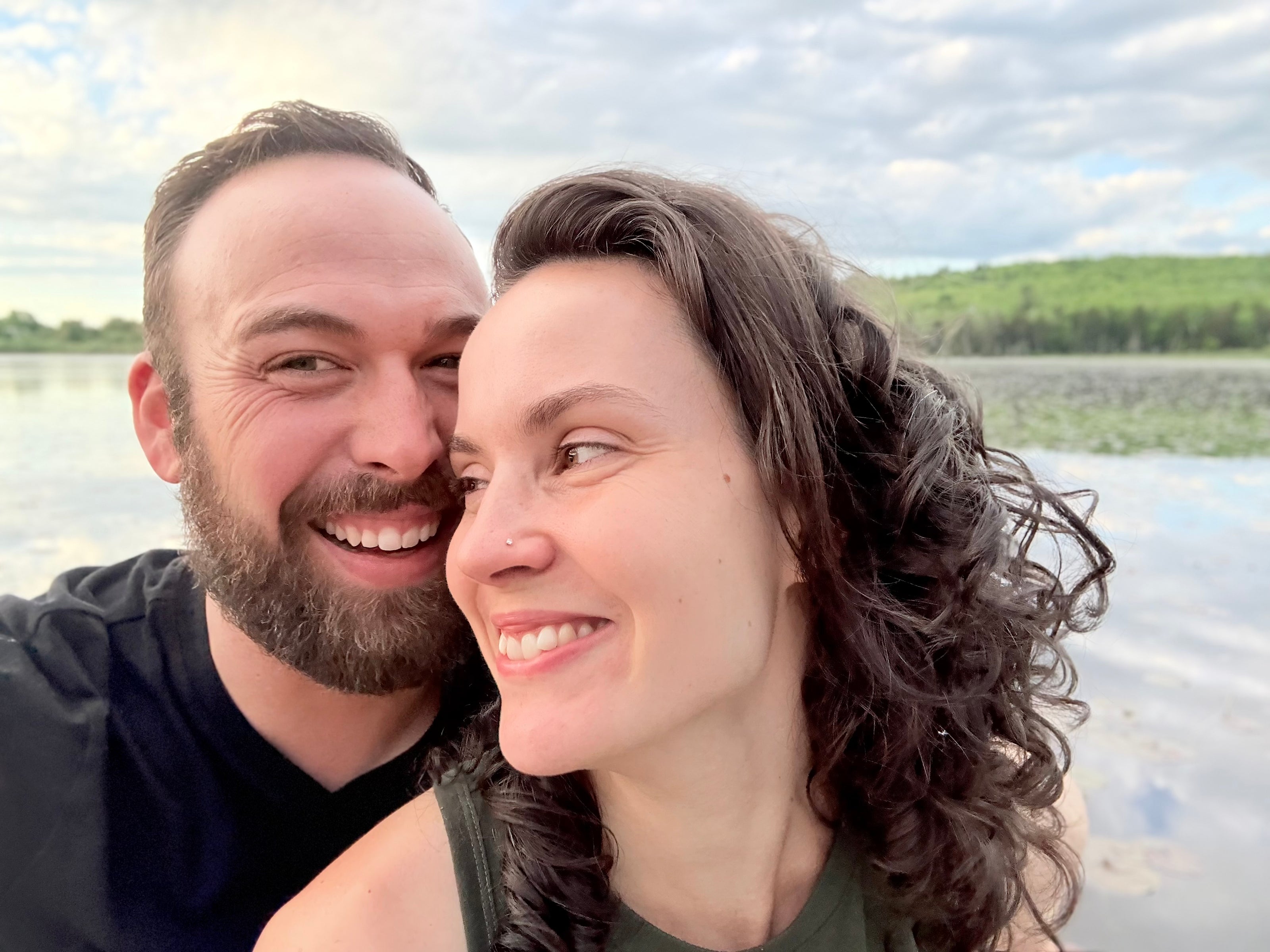 A smiling couple taking a close selfie outdoors by a lake, with a cloudy sky and green shoreline in the background.