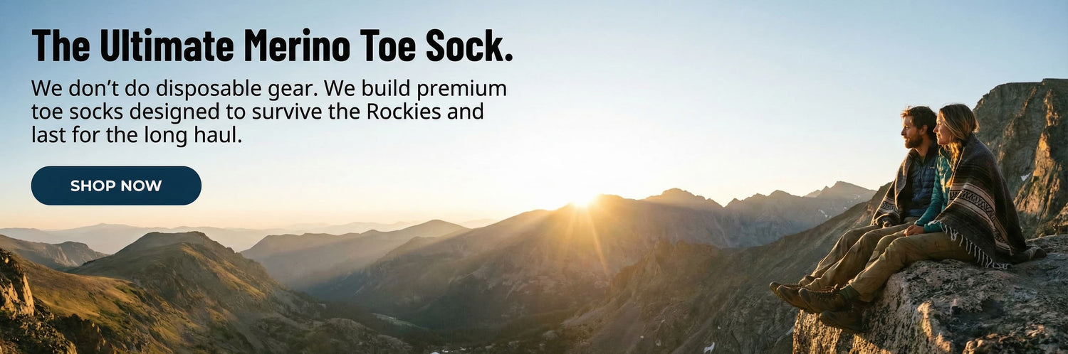 Two people sitting on a mountain ledge with a scenic view of mountains and a lake, promoting merino toe socks.